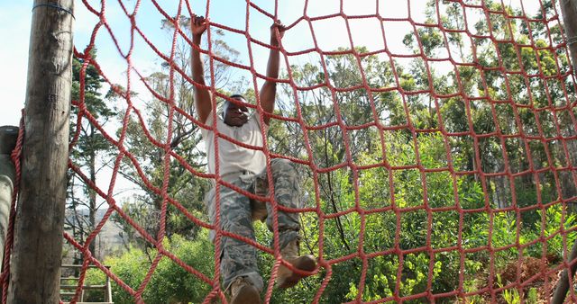 An African American young man is actively climbing a red rope net in an outdoor adventure park, with copy space. His focused expression and dynamic posture convey determination and the enjoyment of physical challenges.