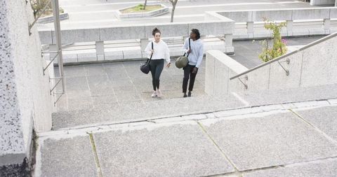 Diverse friends walking up concrete stairs in urban plaza carrying duffel bags and talking