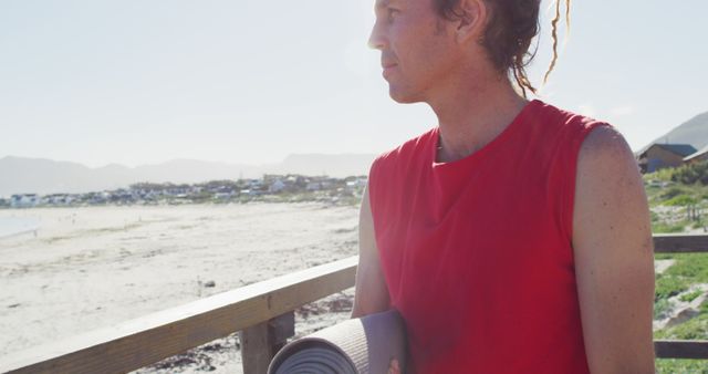 A woman with dreadlocks wearing red shirt holds a yoga mat while standing on a beachside boardwalk. Background includes sandy beach, coastline with people, and houses or cabins in the distance. Perfect for themes related to outdoor fitness, health and wellness, and coastal living.