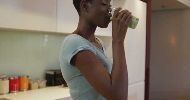 This visual captures a woman enjoying a nutritious green smoothie in a contemporary kitchen. Ideal for illustrating concepts related to healthy living, wellness, morning routines, and home life. Useful for blogs, articles on nutrition, and advertisements promoting healthy eating.