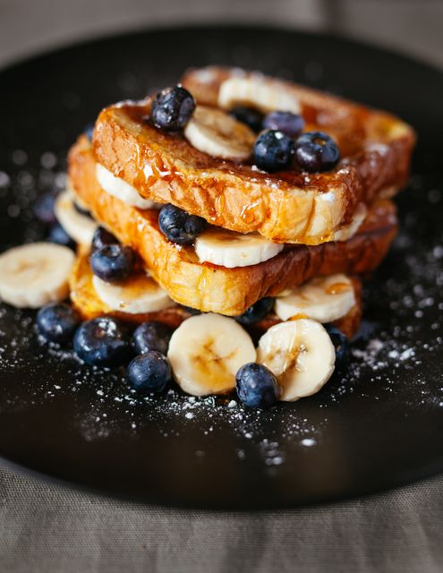 French toast served with sliced bananas and fresh blueberries on black plate. Topped with syrup and powdered sugar, creating a delicious and visually appealing breakfast meal. Ideal for food blogs, recipe websites, breakfast menus, and social media posts for brunch ideas.