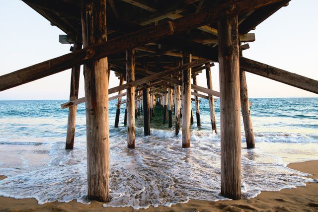 Waves gently hitting the shore underneath a wooden pier at sunset offers a tranquil and scenic view. Ideal for travel brochures, coastal lifestyle illustrations, and nature-themed artworks. The perspective from beneath the pier provides an interesting linear composition, useful for adding depth and focus to design projects.