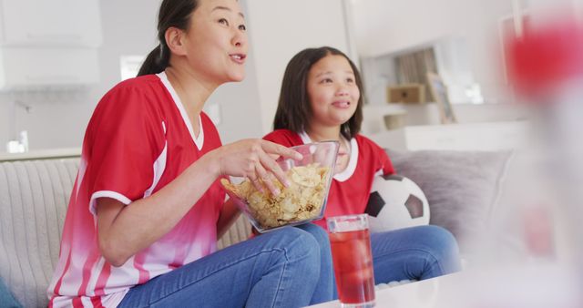 Mother and daughter enjoying a soccer match at home, both wearing matching team jerseys, showcasing family bonding and fandom. The daughter holds a soccer ball while they sit on the couch, focused on the game. Suitable for themes like family activities, sports enthusiasm, and home leisure.