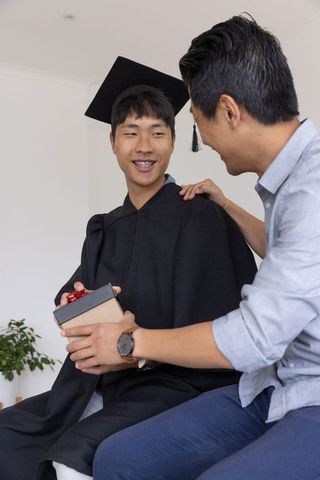 Asian father and son celebrating graduation at home, son wearing cap and gown holding gift
