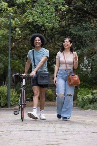 Laughing diverse couple walking in park with bicycle and coffee, casual urban lifestyle