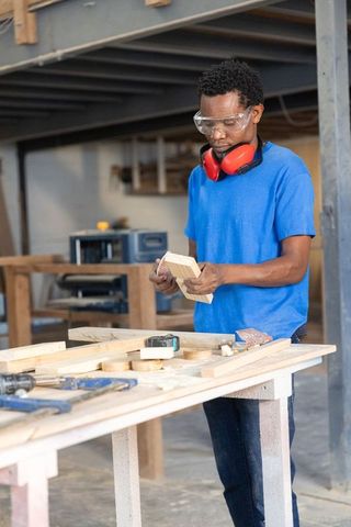 African american carpenter inspecting wooden block on workbench wearing goggles ear protectors