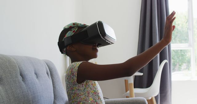African american girl gesturing while using vr headset sitting on the couch at home. family, togetherness and happiness concept