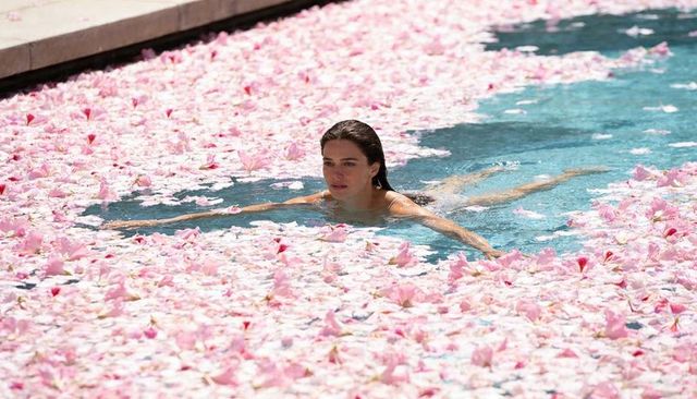 Woman swimming through pink flower petals in sunlit outdoor pool spa luxury relaxation