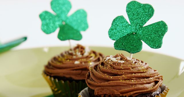 Chocolate cupcakes are adorned with sparkling green shamrock decorations, suggesting a festive treat for St. Patrick's Day celebrations. Their glittering toppers add a touch of whimsy and seasonal flair to the dessert presentation.