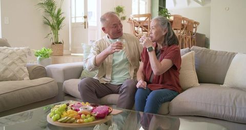 Senior asian couple sipping water and sharing fruit on cozy sunlit living room sofa