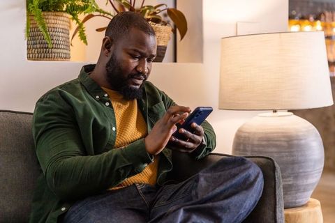 African american man relaxing on sofa using smartphone in cozy modern living room