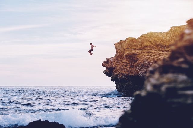 Person enjoying an adrenaline-fueled activity by leaping off a rocky cliff into the ocean below at sunset. Ideal for travel blogs, adventure tourism marketing, and motivational content related to facing fears and seeking thrills.