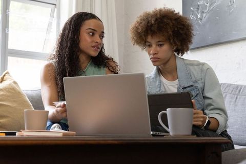 Two women collaborating on laptop and tablet on couch at home for remote work and study