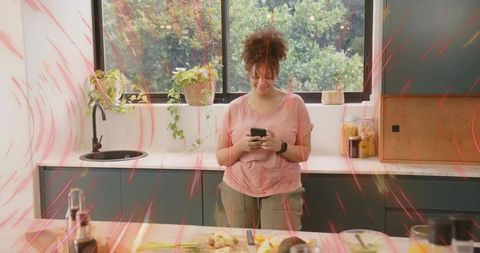 Checking smartphone while prepping ingredients in sunlit modern kitchen with plants