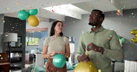 Couple celebrating with metallic teal and gold balloons, confetti falling in living room
