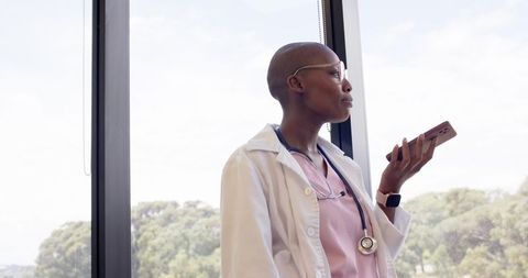 Standing clinician holding smartphone and stethoscope by window, healthcare professional