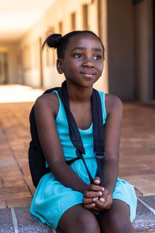African american girl sitting in school corridor wearing turquoise dress and backpack