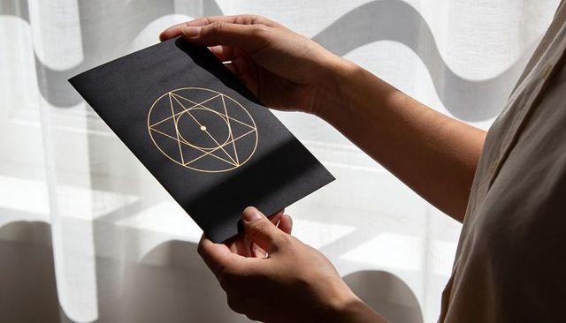 Hands holding black card with gold geometric emblem near sunlit sheer curtains