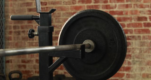 A barbell loaded with weight plates rests on a squat rack in a gym with a brick wall background, with copy space. Fitness enthusiasts use such equipment for strength training and muscle building exercises.