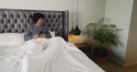 African American man relaxing in bed using smartphone under tufted headboard with plant