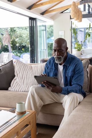 Senior man sitting on sectional sofa using tablet, relaxing in bright modern living room