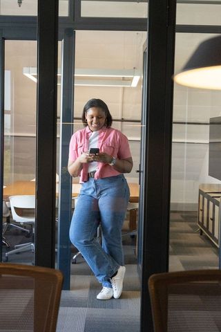 African american woman leaning on glass checking smartphone and smartwatch in modern office