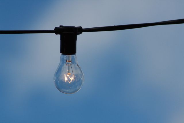 Single light bulb effortlessly hanging on a black wire against a clear blue sky. Ideal for illustrating themes of simplicity, energy, outdoor decoration, or minimalism. Suitable for use in articles about electricity, energy saving, outdoor events, or decoration tips.