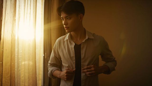 Young asian man buttoning shirt by sunlit window during golden hour contemplative mood