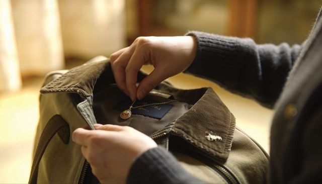 Hands placing vintage locket inside canvas jacket with corduroy collar in warm light