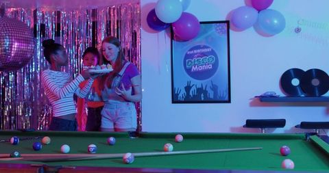 Celebrating 21st disco birthday with diverse friends holding cake near neon pool table