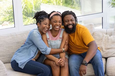 African parents hugging teenage daughter on sofa laughing and bonding in living room