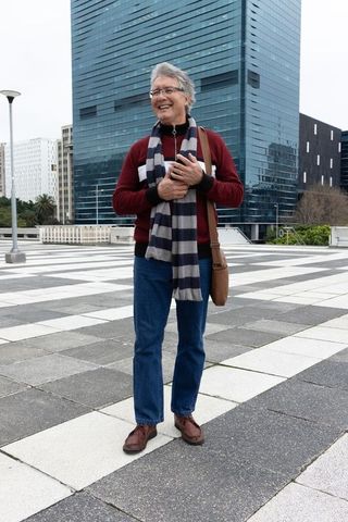 Senior man standing in urban plaza holding smartphone and bag by glass skyscraper
