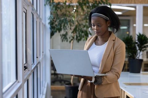 African american professional standing by window holding laptop in modern office