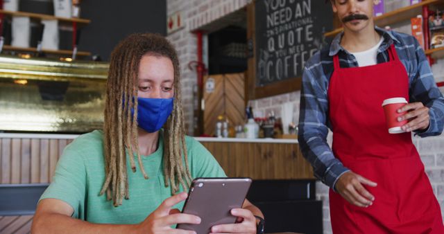 Customer seated in coffee shop using tablet, wearing face mask for safety. Barista in red apron approaching with drink. Ideal for concepts of technology in service industry, protective health measures, cafe environment.