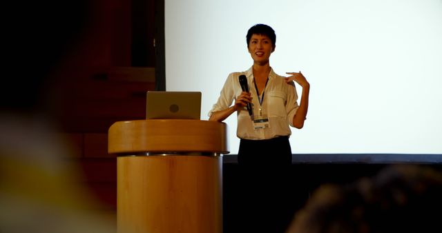 Front view of beautiful young Asian businesswoman speaking in business seminar in auditorium. She is holding microphone 4k