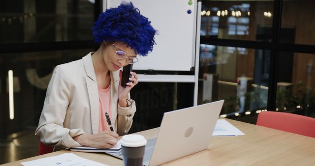 A professional woman with striking blue hair at a modern office desk. She is talking on the phone while working on documents with a laptop open beside her. A coffee cup is placed on the table, hinting at a busy workday. The setting suggests a creative and contemporary work environment, possibly ideal for artwork or office culture content. Perfect for imagery focusing on modern business practices, unique office settings, or promotional material for workplace diversity and inclusiveness.