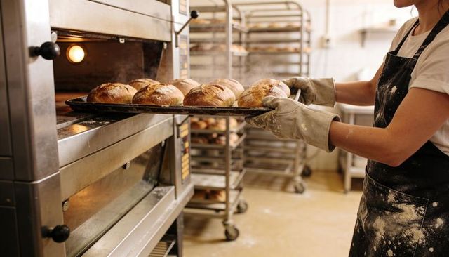 Baker removing fresh sourdough loaves from deck oven in busy artisan bakery kitchen