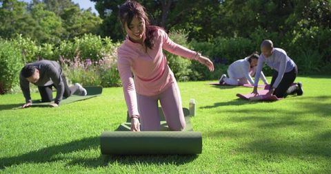 Diverse friends preparing outdoor yoga, rolling mats on sunlit park lawn