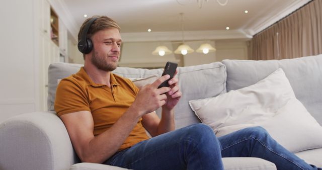 Portrait of happy caucasian man sitting on sofa wearing headphones and using smartphone. staying at home in isolation during quarantine lockdown.