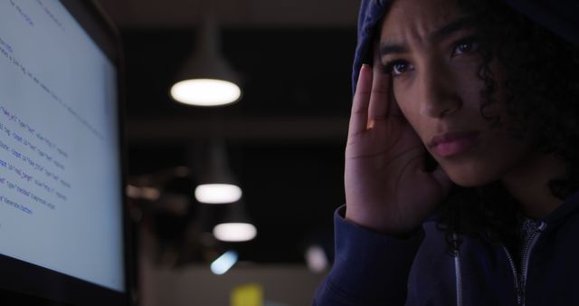 Young biracial woman looks concerned at a computer screen, with copy space. She's facing a challenging task or problem in a dimly lit office or home.