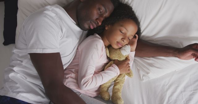 Father and daughter lying together on bed in intimate cozy moment. Daughter is holding a teddy bear. Depicts strong family bond and affection. Ideal for use in advertisements, family care services promotions, parenting blogs, and articles on family well-being and relaxation.