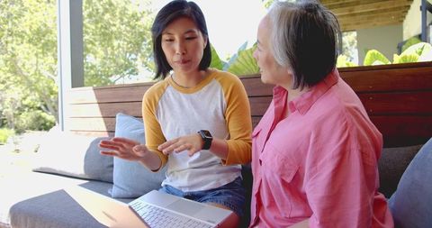 Asian mother and adult daughter sitting on sunlit patio bench collaborating on laptop
