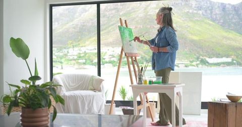 Mature woman painting on easel in sunlit coastal studio with mountain and sea view