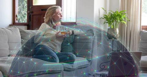 Mature woman relaxing with mug on couch while remote working with digital network overlay