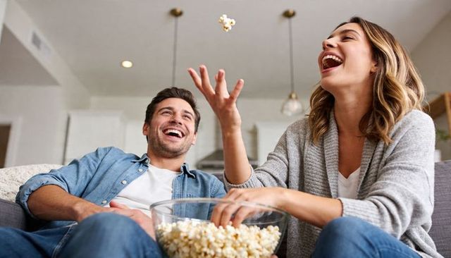 Laughing couple sharing popcorn on couch in modern open-plan living room, cozy date night