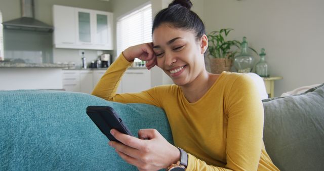 Woman in casual yellow sweater sitting on sofa and using smartphone in modern living room. Ideal for use in advertisements promoting smart gadgets, apps, or happy home living concepts.