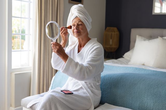 Smiling mature caucasian woman holding mirror brushing blusher on face in bedroom, copy space. Self care, health, beauty and senior lifestyle concept.