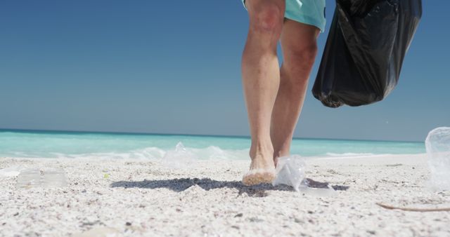 Legs of caucasian man with black bag cleaning beach of plastic on beach by sea, copy space. Recycling, lifestyle, vacation, summer concept, unaltered.