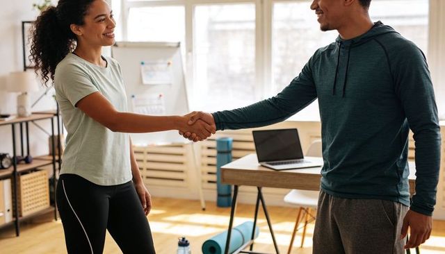 Fitness coach meeting client shaking hands in bright home studio with yoga gear and laptop
