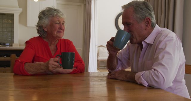 Senior couple enjoying coffee together at home, sitting at wooden table and engaging in conversation. Suitable for themes related to senior life, retirement, family moments, and health and wellness advertisements.
